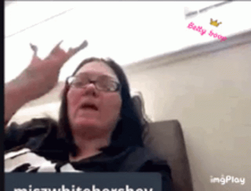 A Woman Wearing Glasses Is Sitting On A Couch And Making A Devil Horns Gesture . GIF
