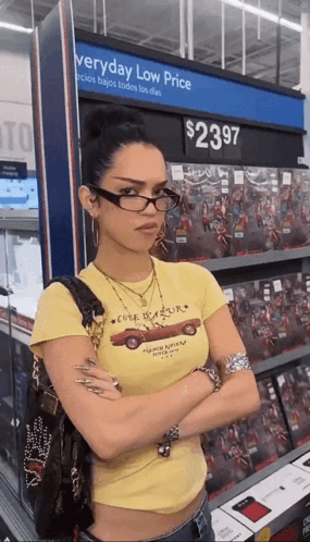 A Woman Wearing Glasses Is Standing In Front Of A Sign That Says Everyday Low Price GIF