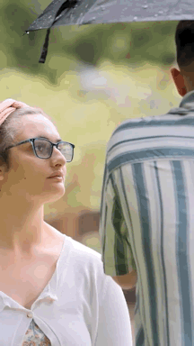 A Woman Wearing Glasses Is Standing Next To A Man Holding An Umbrella Over Her Head . GIF
