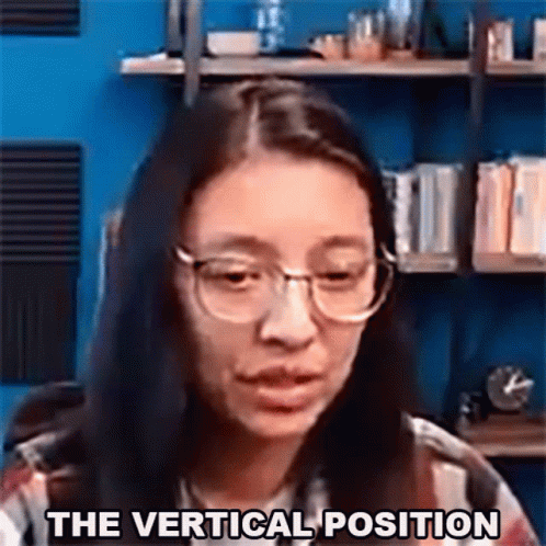 A Woman Wearing Glasses Says The Vertical Position In Front Of A Bookshelf . GIF