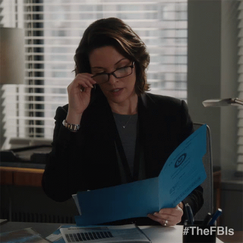A Woman Wearing Glasses Sits At A Desk Holding A Blue Folder With The Fbi Logo GIF