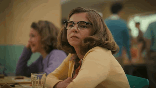 A Woman Wearing Glasses Sits At A Table With A Glass Of Water In Front Of Her GIF