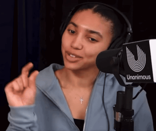 A Woman Wearing Headphones And A Cross Necklace Stands In Front Of A Unanimous Microphone GIF
