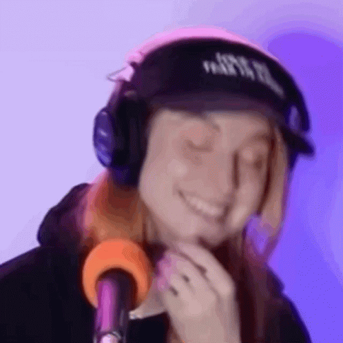 A Woman Wearing Headphones And A Hat Is Smiling And Looking At The Camera . GIF