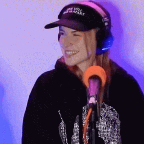 A Woman Wearing Headphones And A Hat Is Standing In Front Of A Microphone . GIF