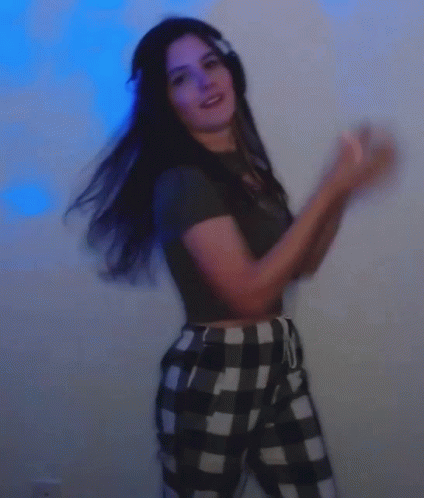 A Woman Wearing Headphones And Plaid Pants Is Dancing In Front Of A Blue Wall . GIF