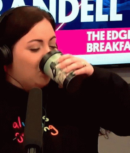 A Woman Wearing Headphones Drinks From A Cup In Front Of A Screen That Says " The Edge Breakfast " GIF