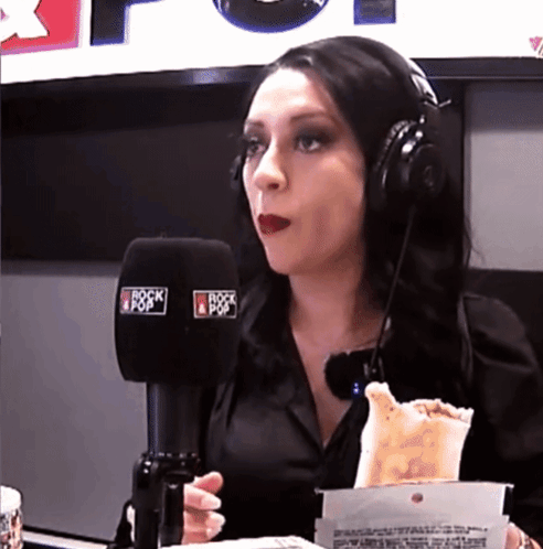 A Woman Wearing Headphones Is Eating A Taco In Front Of A Microphone That Says Rock & Pop GIF