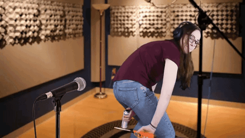 A Woman Wearing Headphones Is Kneeling In Front Of A Microphone And Holding A Can Of Elmer 'S Glue GIF