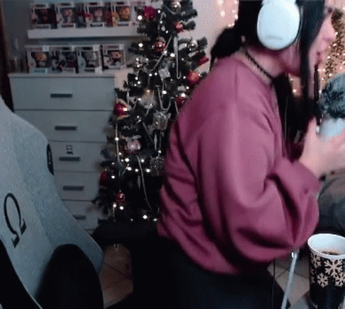 A Woman Wearing Headphones Is Sitting In Front Of A Christmas Tree Holding A Cup Of Coffee . GIF