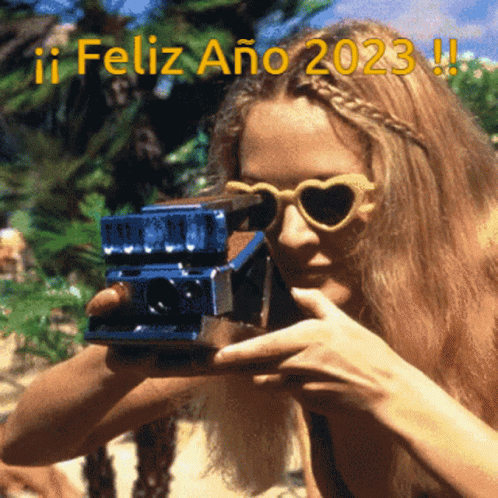 A Woman Wearing Heart Shaped Sunglasses Is Taking A Picture With A Polaroid Camera With The Words Feliz Ano 2023 Written GIF