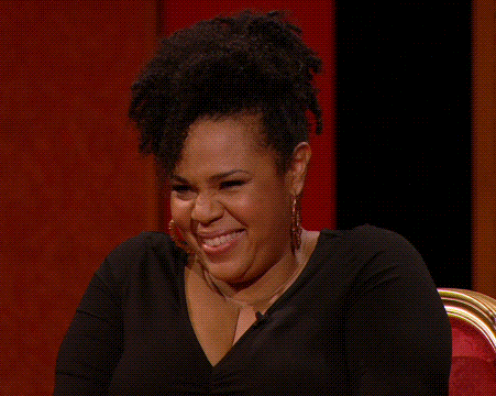 A Woman Wearing Hoop Earrings And A Black Shirt Is Laughing While Sitting In A Chair . GIF