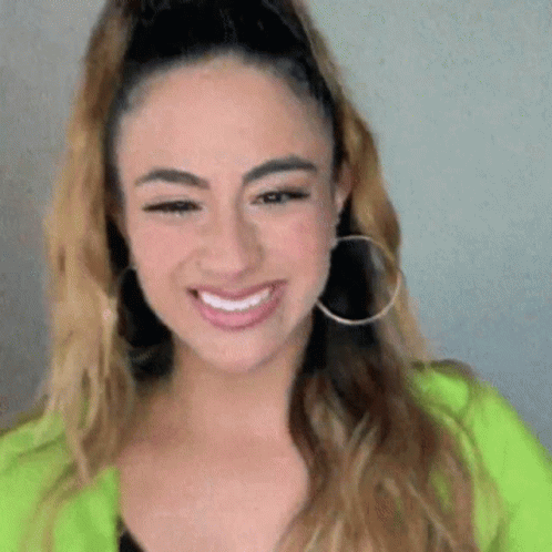 A Woman Wearing Hoop Earrings And A Green Jacket Is Smiling . GIF