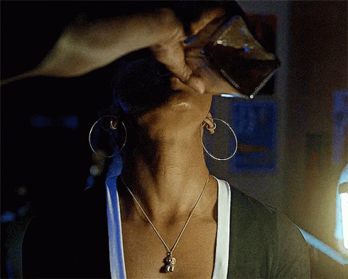 A Woman Wearing Hoop Earrings And A Necklace With A Monkey Pendant Is Drinking From A Glass GIF