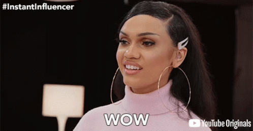 A Woman Wearing Hoop Earrings And A Pink Turtleneck Sweater Is Saying Wow GIF