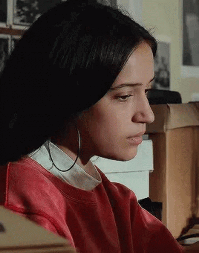 A Woman Wearing Hoop Earrings And A Red Sweater Is Sitting At A Desk . GIF
