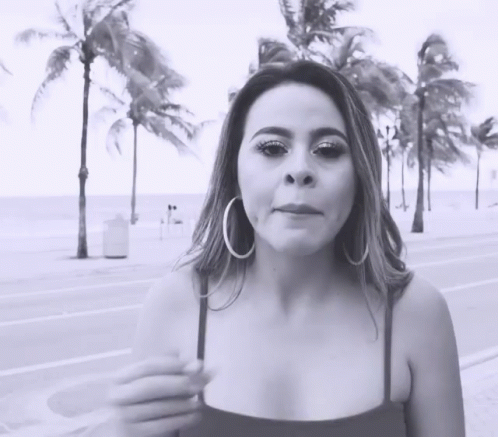 A Woman Wearing Hoop Earrings Is Standing On A Sidewalk In Front Of Palm Trees GIF