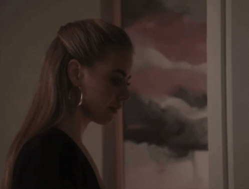 A Woman Wearing Hoop Earrings Stands In Front Of A Framed Picture GIF