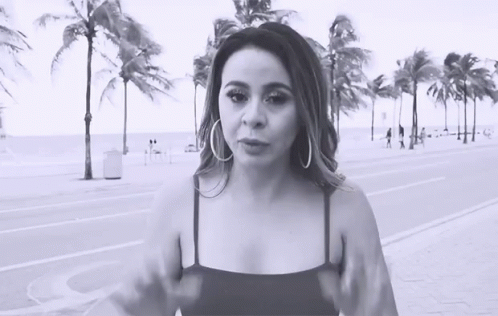 A Woman Wearing Hoop Earrings Stands In Front Of Palm Trees On The Beach GIF