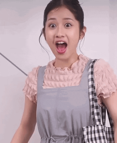 A Woman Wearing Overalls And A Pink Shirt Is Holding A Checkered Bag And Making A Surprised Face GIF