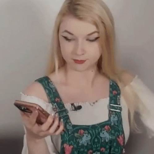 a woman wearing overalls and a white shirt is holding a cell phone .