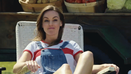 A Woman Wearing Overalls Sits In A Chair With Her Legs Crossed GIF