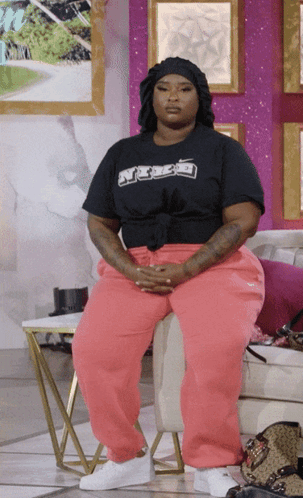 A Woman Wearing Pink Sweatpants And A Black Shirt That Says ' A ' On It Sits In A Chair GIF