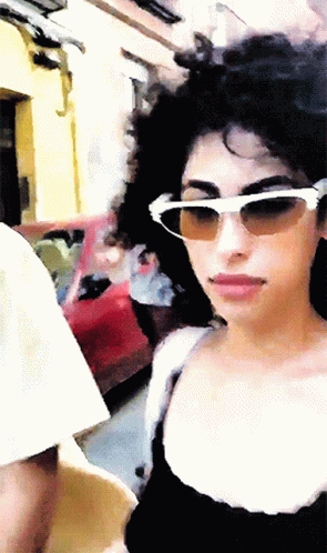 A Woman Wearing Sunglasses And A Black Top Is Walking Down A Street GIF