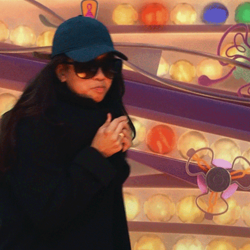 A Woman Wearing Sunglasses And A Blue Hat Stands In Front Of A Display Of Colorful Balls GIF