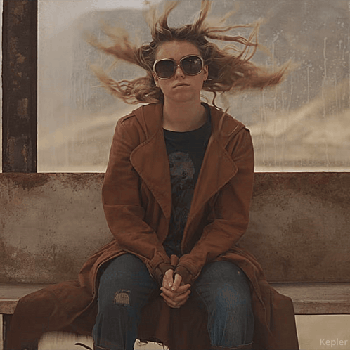 A Woman Wearing Sunglasses And A Brown Coat Sits On A Bench In Front Of A Window That Says Kepler On It GIF