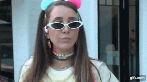 A Woman Wearing Sunglasses And A Colorful Headband Is Standing In Front Of A White Building . GIF
