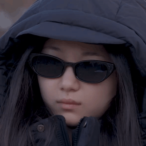 A Woman Wearing Sunglasses And A Hooded Jacket Looks At The Camera GIF
