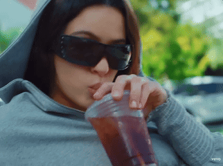 A Woman Wearing Sunglasses And A Hoodie Drinks From A Plastic Cup GIF