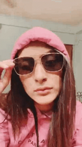 A Woman Wearing Sunglasses And A Pink Hoodie Is Taking Off Her Sunglasses . GIF