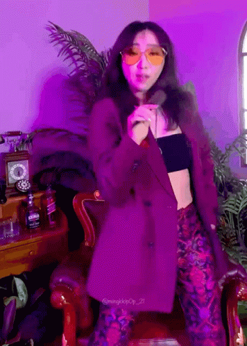 A Woman Wearing Sunglasses And A Purple Jacket Stands In Front Of A Purple Wall GIF
