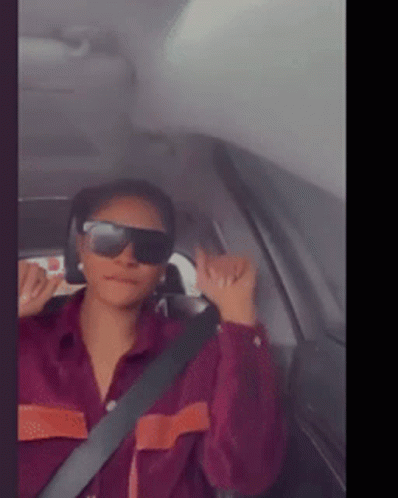 A Woman Wearing Sunglasses And A Purple Shirt Is Sitting In A Car GIF