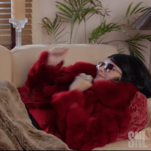 A Woman Wearing Sunglasses And A Red Fur Coat Is Laying On A Couch With Snl Written On It GIF