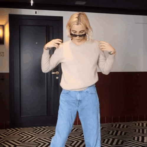 A Woman Wearing Sunglasses And A Sweater Is Standing In Front Of A Door That Has The Number 23 On It GIF