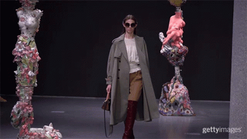 A Woman Wearing Sunglasses And A Trench Coat Walks Down A Runway In Front Of Sculptures GIF