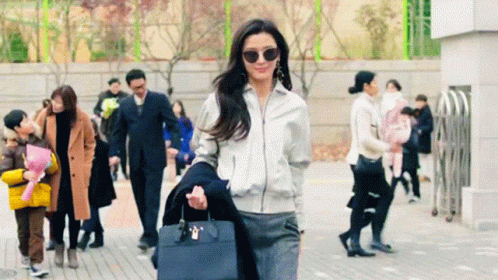 A Woman Wearing Sunglasses And A White Jacket Is Walking Down A Sidewalk GIF
