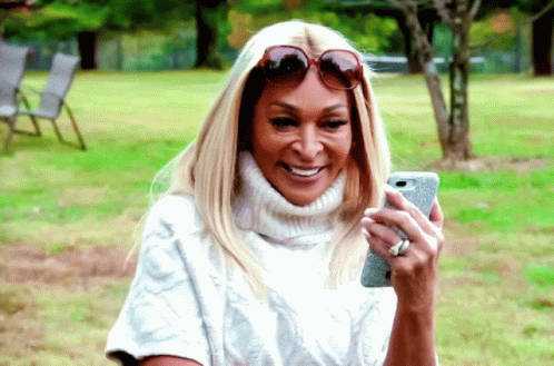 A Woman Wearing Sunglasses And A White Sweater Is Looking At Her Phone GIF