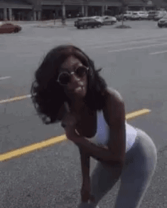 A Woman Wearing Sunglasses And A White Tank Top Is Standing On The Side Of The Road . GIF