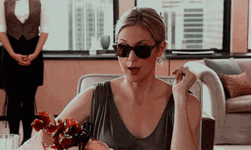 A Woman Wearing Sunglasses And Earrings Sits At A Table GIF