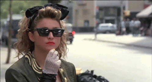 A Woman Wearing Sunglasses And Gloves Is Standing On A City Street GIF