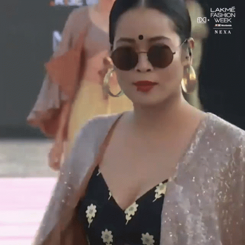 A Woman Wearing Sunglasses And Hoop Earrings Is Walking Down A Runway At Lakme Fashion Week GIF
