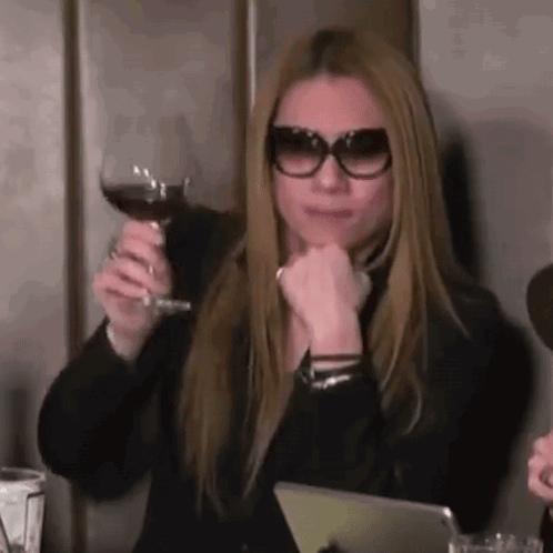 A Woman Wearing Sunglasses Is Sitting At A Table Holding A Glass Of Red Wine . GIF