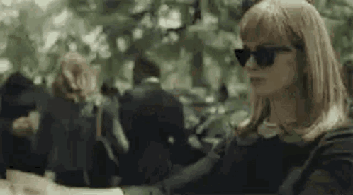 A Woman Wearing Sunglasses Is Sitting At A Table In A Crowded Park . GIF