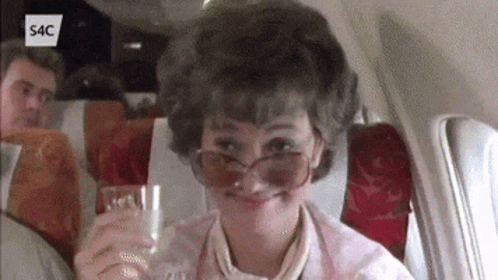 A Woman Wearing Sunglasses Is Sitting On An Airplane Holding A Glass GIF