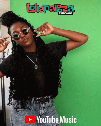 A Woman Wearing Sunglasses Stands In Front Of A Green Screen That Says ' Lolapaloza Chicago ' On It GIF