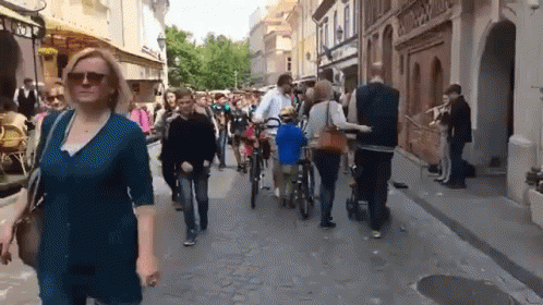 A Woman Wearing Sunglasses Walks Down A Busy Street With People Walking And Riding Bikes GIF